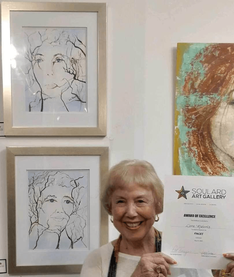 Lora Roberts smiling and holding an Award of Excellence certificate from Soulard Art Gallery's FACES exhibition, standing beside two of her framed ink-and-watercolor portraits of faces with tree-branch hair.
