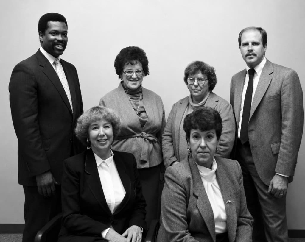 Black and white office group photograph of six professionally dressed people, two seated in front and four standing behind.