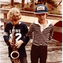 Two boys outdoors, one playing a saxophone in a Lions 42 shirt, the other in a striped shirt and cap.