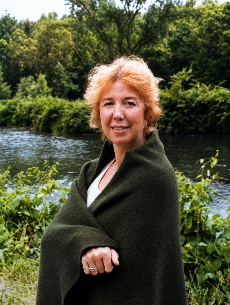 Portrait of a woman with red hair wrapped in a dark green blanket standing beside a tree-lined river.