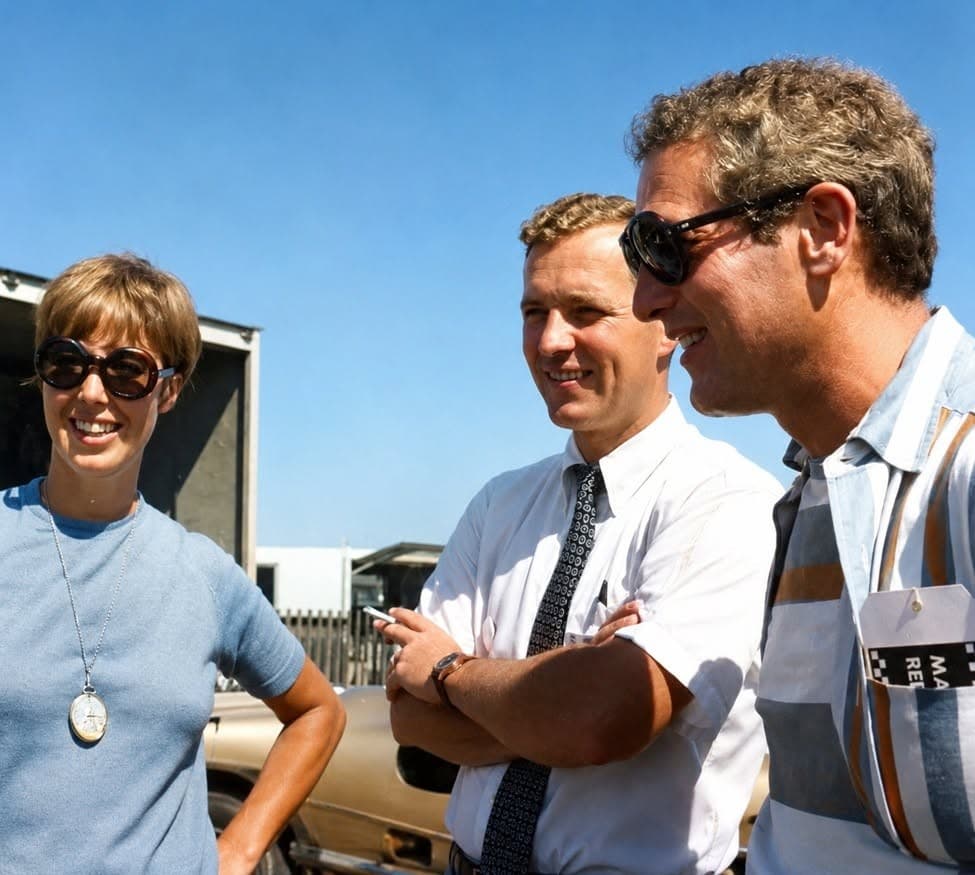 Vintage color photograph of a smiling woman in round sunglasses and a pocket-watch necklace talking with two men beside a race car and transporter under a blue sky.